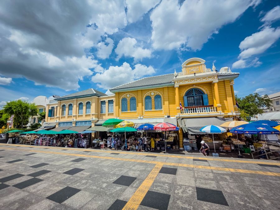 Marché animé devant bâtiment colonial jaune sous ciel nuageux